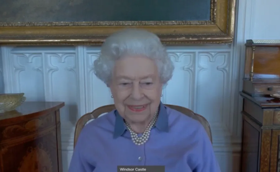 Elderly woman with white hair in a blue blouse and pearl necklace, seated indoors, smiling.