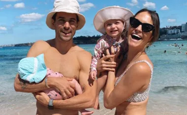A happy couple with two children on a sunny beach; both adults wearing hats and swimsuits, one child cradled, another held.