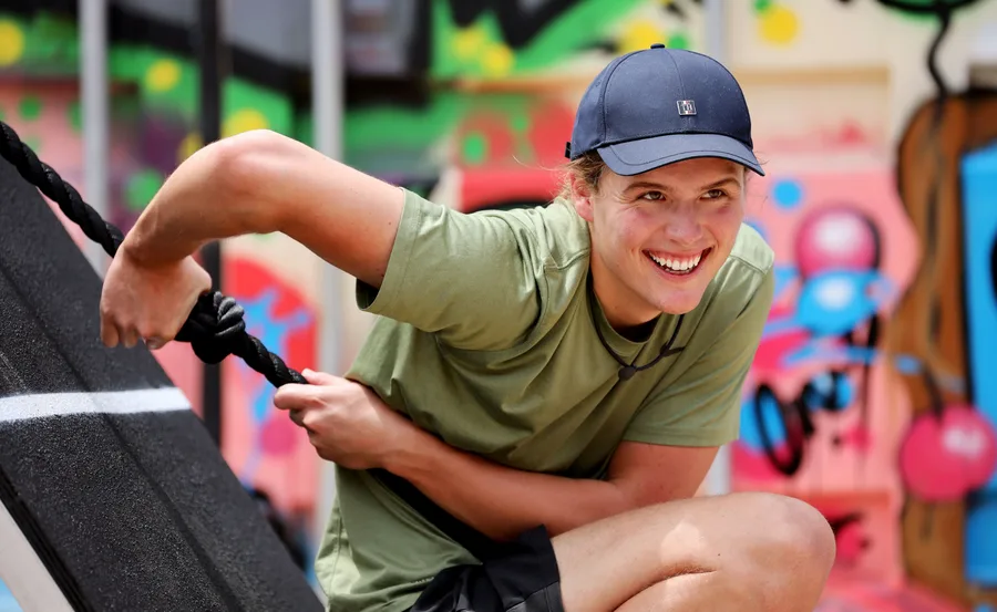 Young person in a cap smiles while climbing a rope with a colorful graffiti background.
