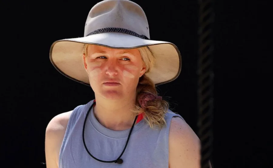 A woman in a wide-brimmed hat with a braid and face paint stands against a dark background, looking off to the side.