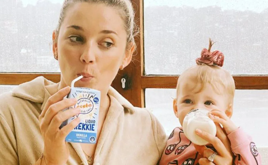 Woman and child drinking milk, woman with a beverage carton and child with a baby bottle, smiling together indoors.
