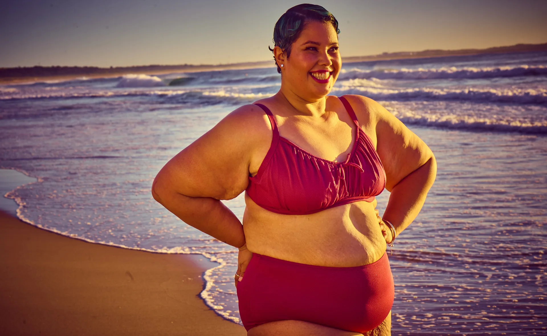 A woman in a red bikini smiles confidently on a sunny beach with waves in the background.