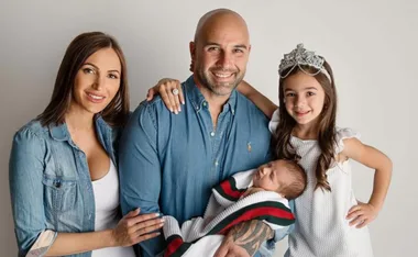 A smiling family of four, including parents, a young girl with a crown, and a baby wrapped in a white blanket.