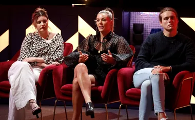Three people seated in red chairs on a stage, engaged in a discussion.