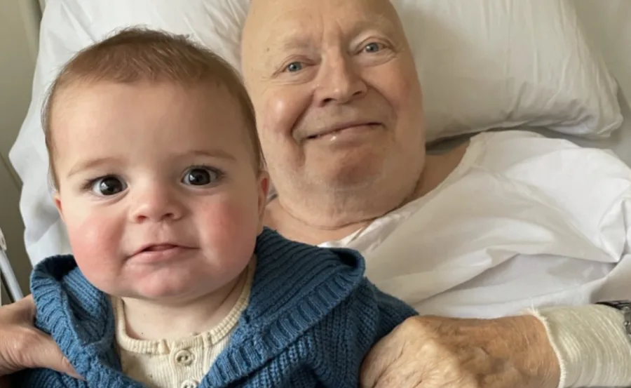 Elderly man in bed holds a baby wearing a blue sweater, both smiling at the camera.