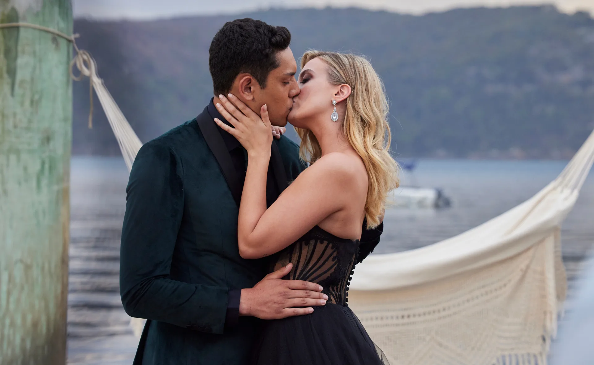 A couple kisses passionately by a lakeside, with a hammock and blurred background.