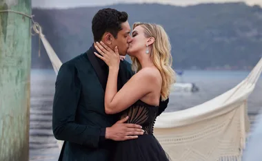 A couple kisses passionately by a lakeside, with a hammock and blurred background.