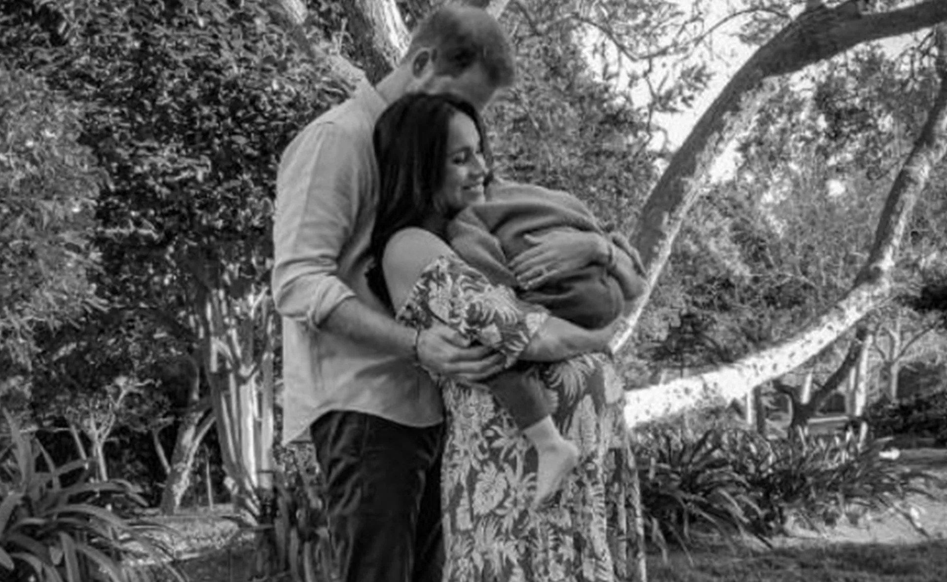 A couple standing in a garden, holding a baby wrapped in a blanket, sharing a tender moment together.