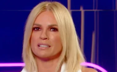 Person with long blonde hair wearing white against a blue background with pink neon light.