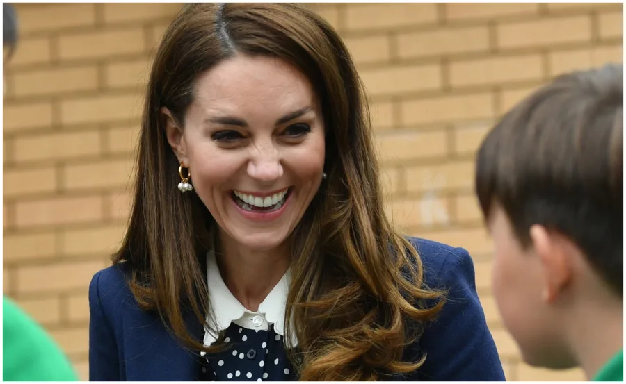 Smiling woman with long brown hair and pearl earrings, engaging with a child, brick wall in the background.