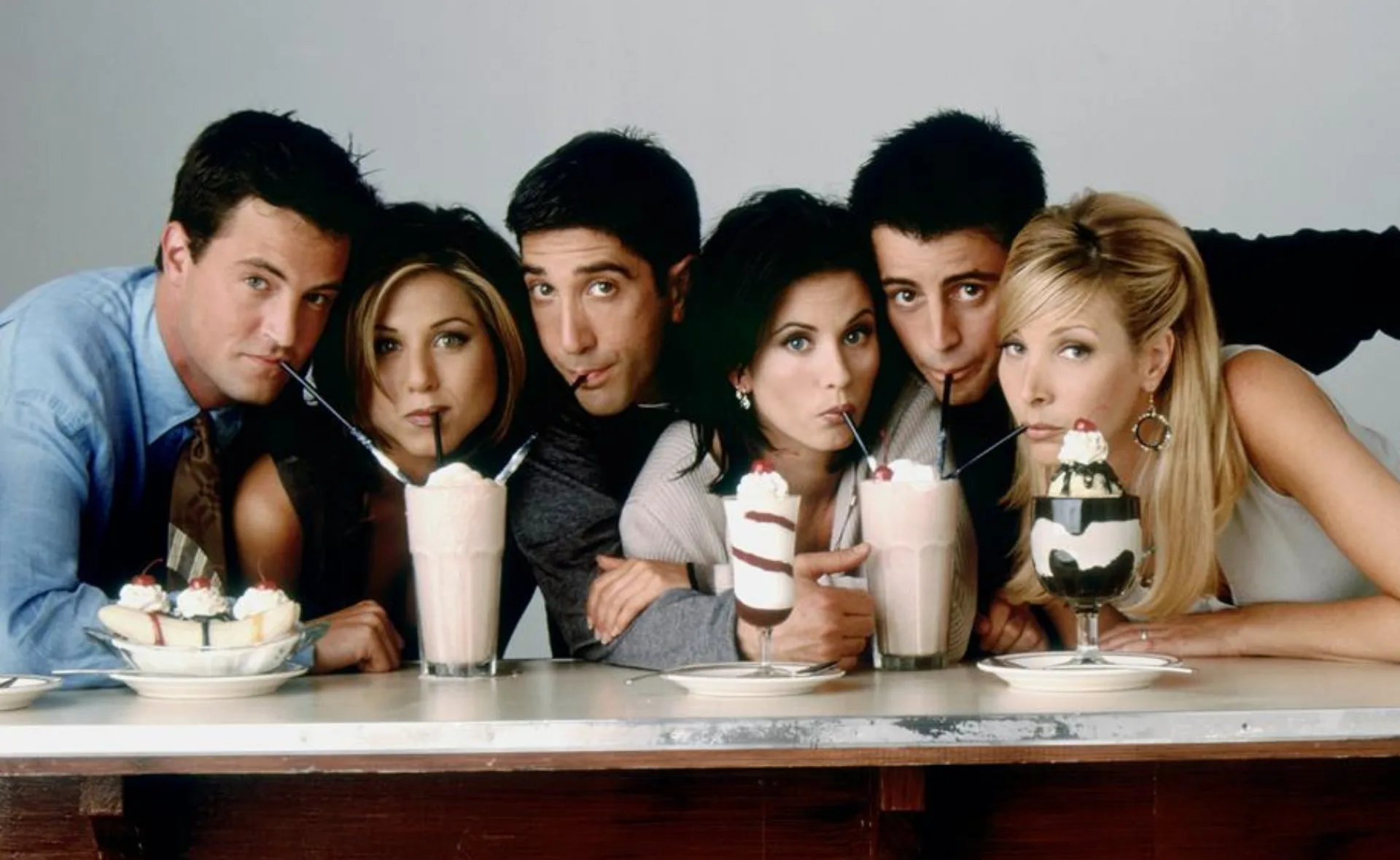 Six friends drinking milkshakes at a table, smiling and leaning close together.