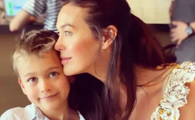 A woman with long dark hair embraces a young boy with short hair, both smiling softly, in a warm indoor setting.