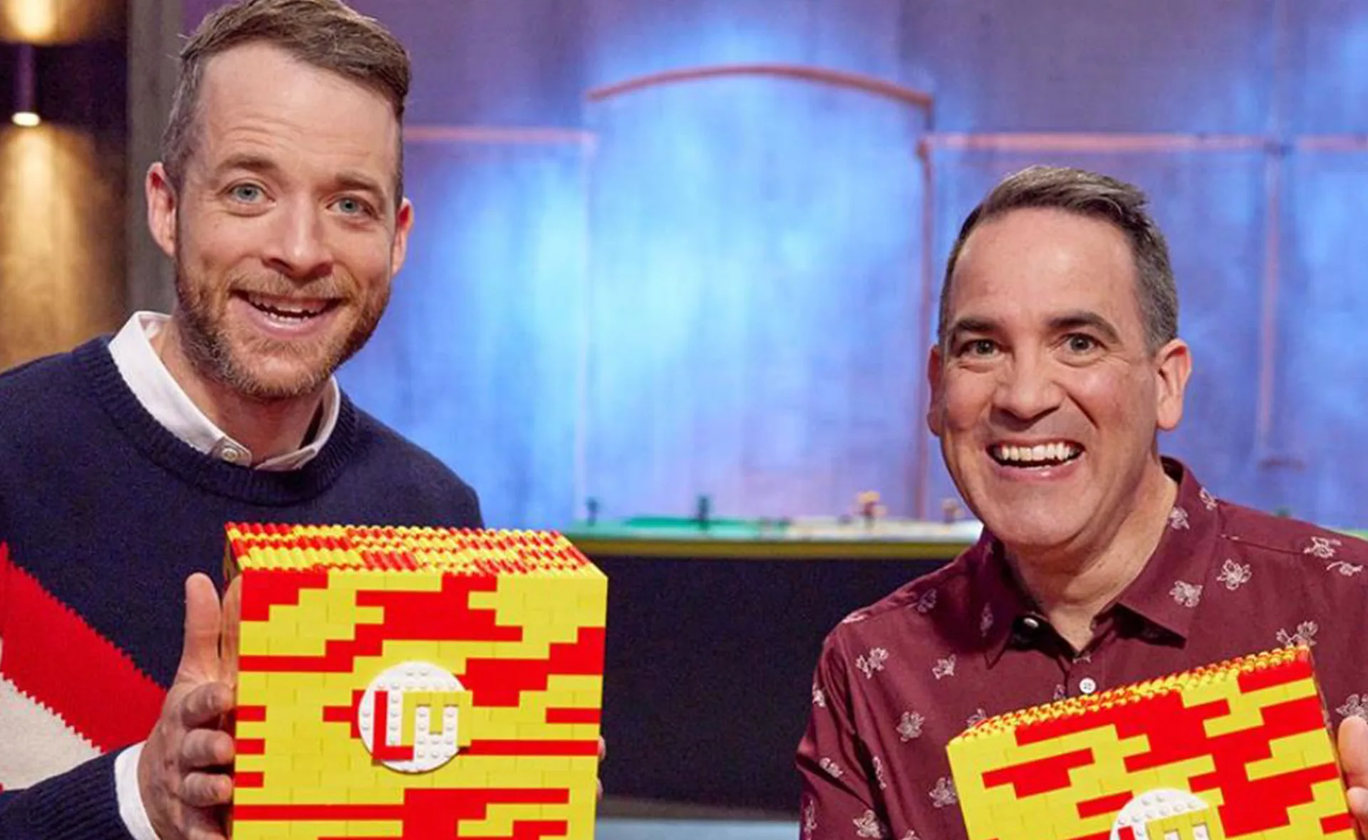 Two smiling individuals holding red and yellow LEGO creations with LM logo on a 'Lego Masters Australia' set.