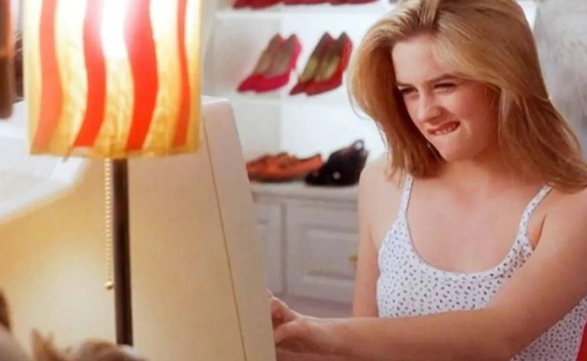 A woman in a white tank top looks at a computer screen with a thoughtful expression, biting her lip.
