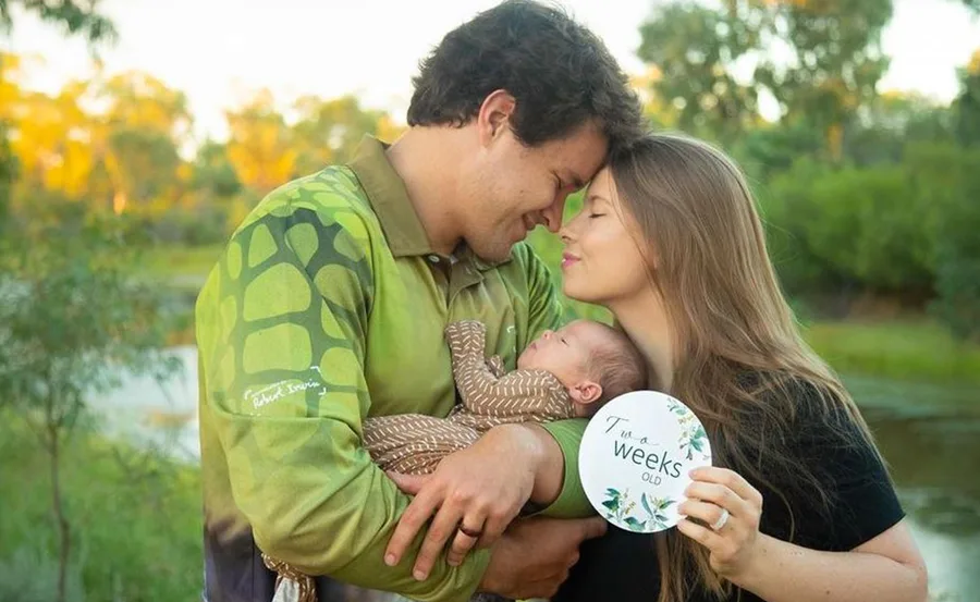 Family cuddling their newborn, who is two weeks old, outdoors with greenery in the background.