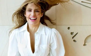 A person with long hair and a white blouse is smiling joyfully, standing in front of a light-colored wall.