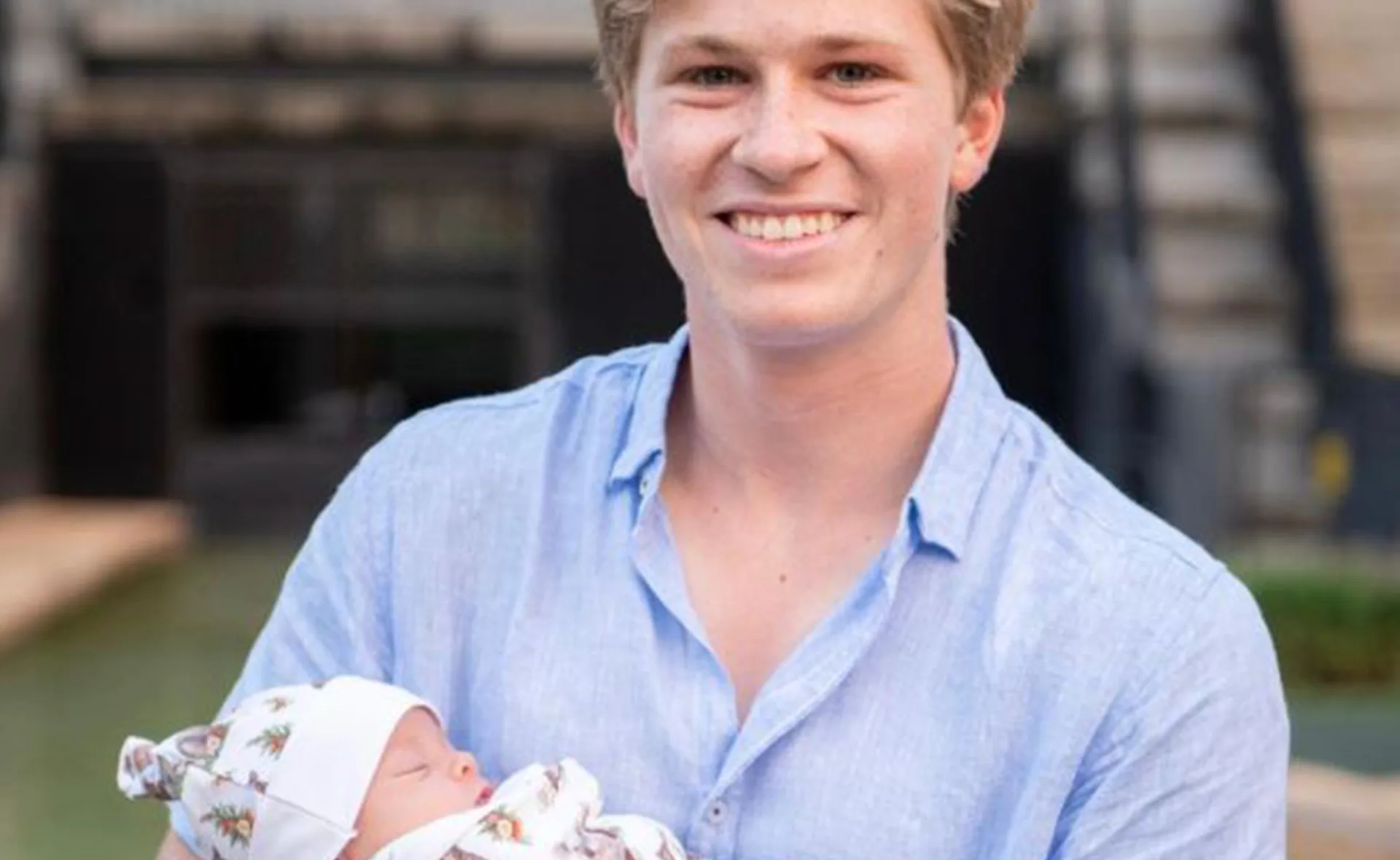 Young man in a blue shirt smiling while holding a baby wrapped in a patterned blanket outdoors.