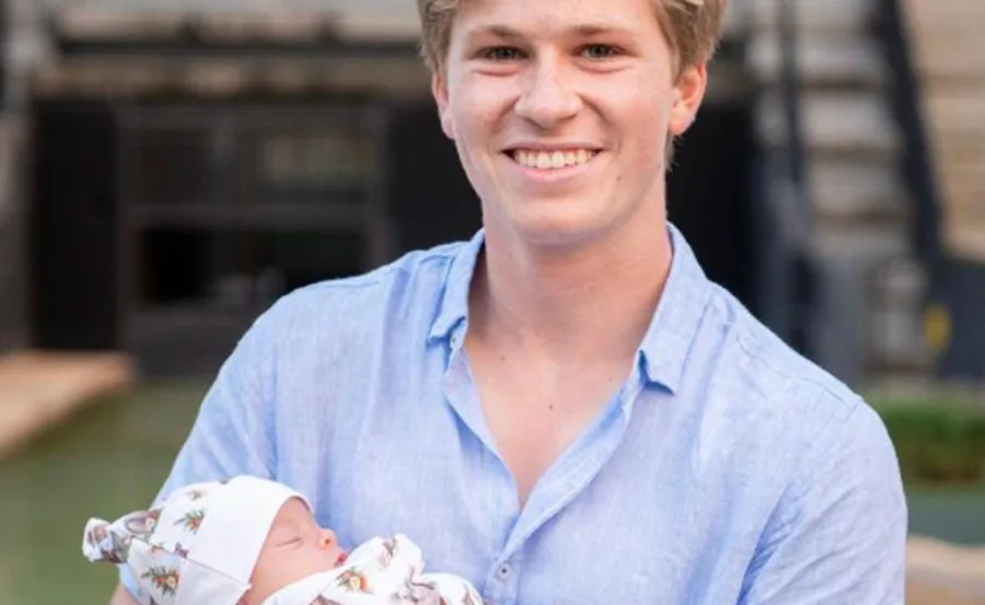 Young man in a blue shirt smiling while holding a baby wrapped in a patterned blanket outdoors.