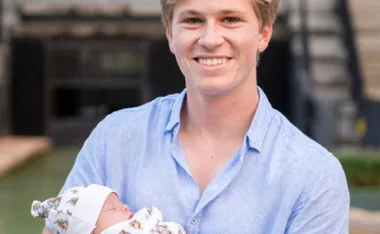 Young man in a blue shirt smiling while holding a baby wrapped in a patterned blanket outdoors.