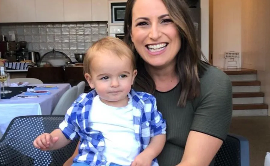 Woman smiling and holding a young child in a blue plaid shirt, seated in a kitchen setting.