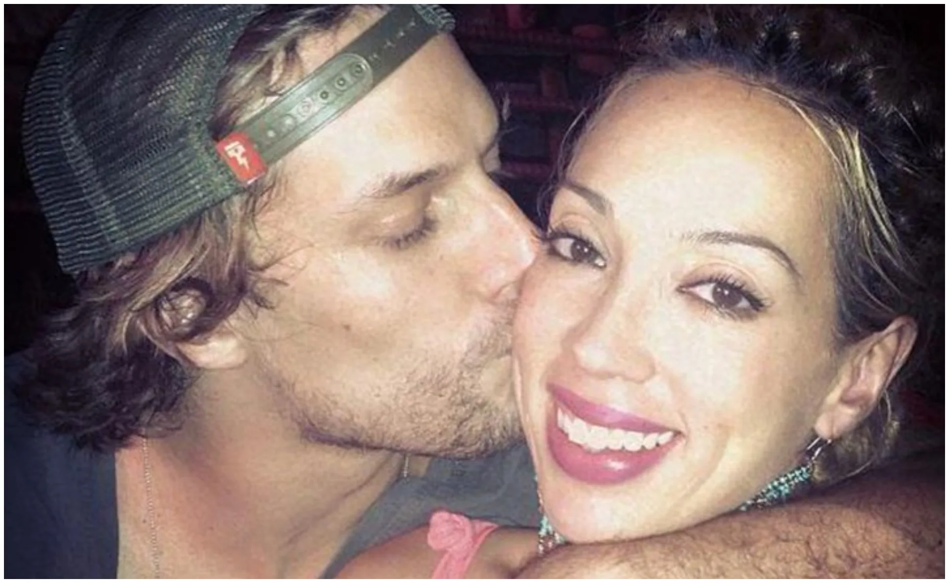 Man kissing woman on cheek; both are smiling. Woman has bright lipstick and earrings; man's eyes are closed, wearing a cap.