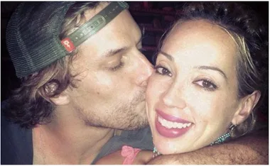 Man kissing woman on cheek; both are smiling. Woman has bright lipstick and earrings; man's eyes are closed, wearing a cap.