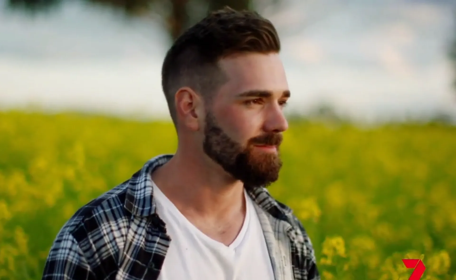 A man with a beard and plaid shirt stands in a field of yellow flowers, looking to the right.
