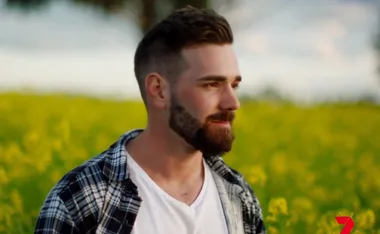 A man with a beard and plaid shirt stands in a field of yellow flowers, looking to the right.
