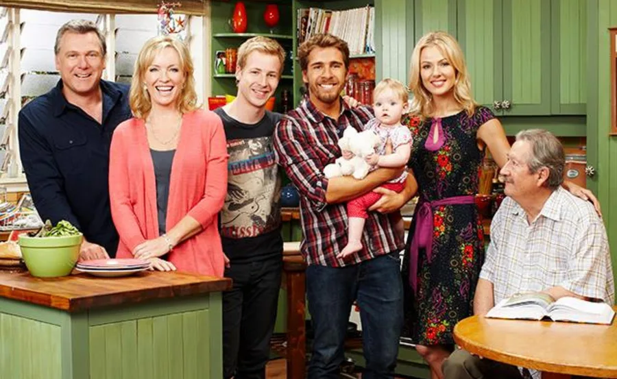 A group of six adults and a baby smiling in a kitchen setting, with green cabinets in the background.