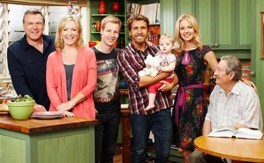 A group of six adults and a baby smiling in a kitchen setting, with green cabinets in the background.