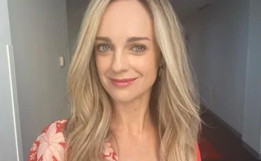 Person with long blonde hair and a floral-patterned top, smiling in a softly lit hallway.