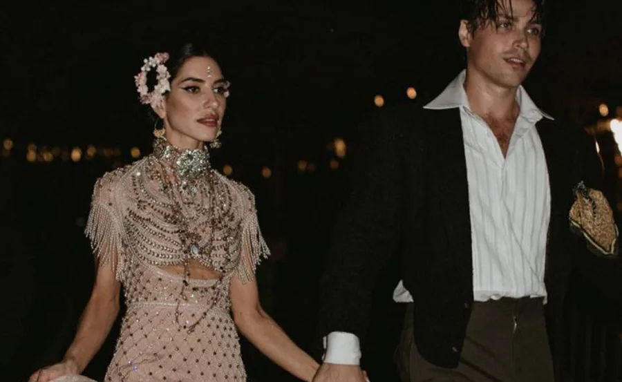 Couple dressed in elegant attire, holding hands, with dark background and ambient lighting.