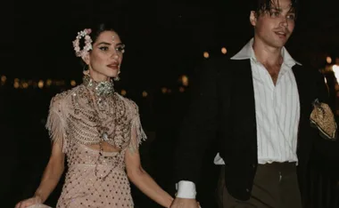 Couple dressed in elegant attire, holding hands, with dark background and ambient lighting.
