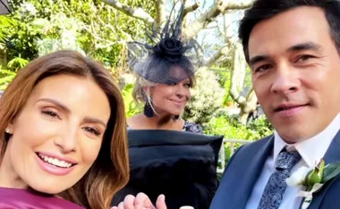 A couple smiling with a woman in a hat behind them, outdoors with trees in the background.