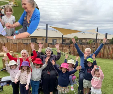 Elderly women and young children with raised arms, smiling outdoors in a garden setting, under cloudy sky.