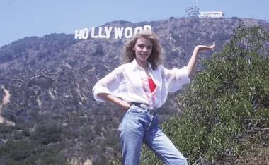 Person posing in front of the Hollywood sign, wearing a white blouse and jeans, with hand raised towards the sign.