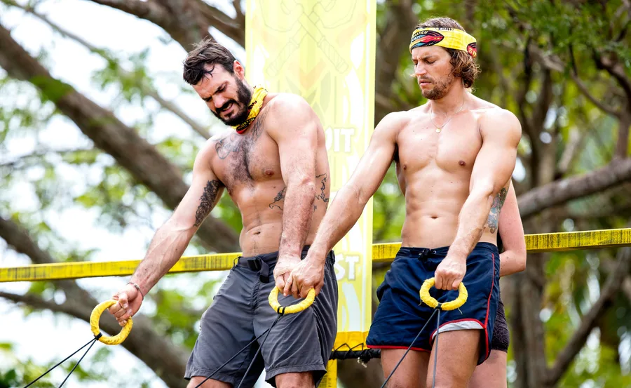 Two men in a challenge, holding ropes, wearing yellow headbands, surrounded by trees.