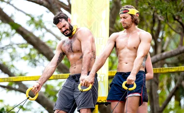 Two men in a challenge, holding ropes, wearing yellow headbands, surrounded by trees.