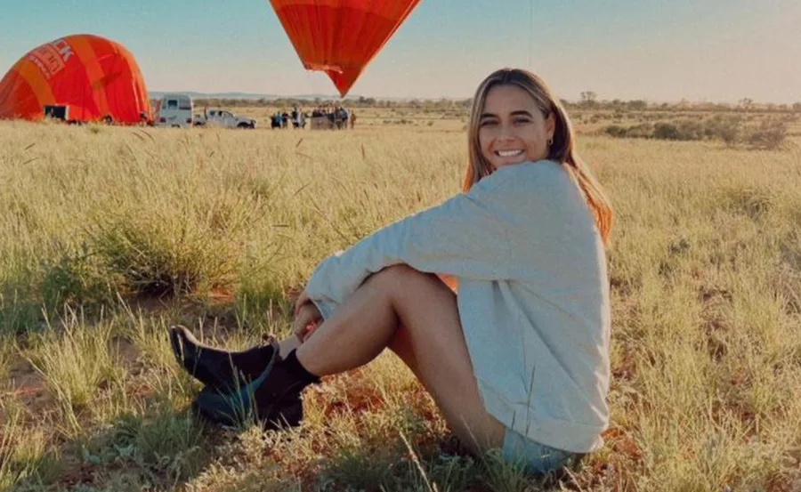 Woman sits on grass with hot air balloons in the background, smiling at the camera.