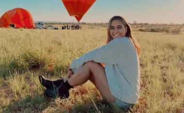 Woman sits on grass with hot air balloons in the background, smiling at the camera.