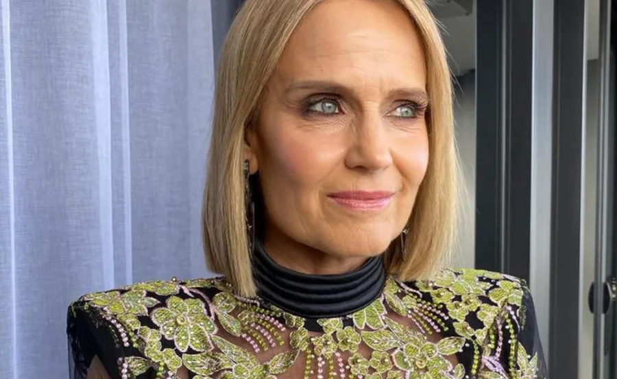 A woman with shoulder-length blonde hair in an elegant, floral embroidered top, standing near a window with white curtains.