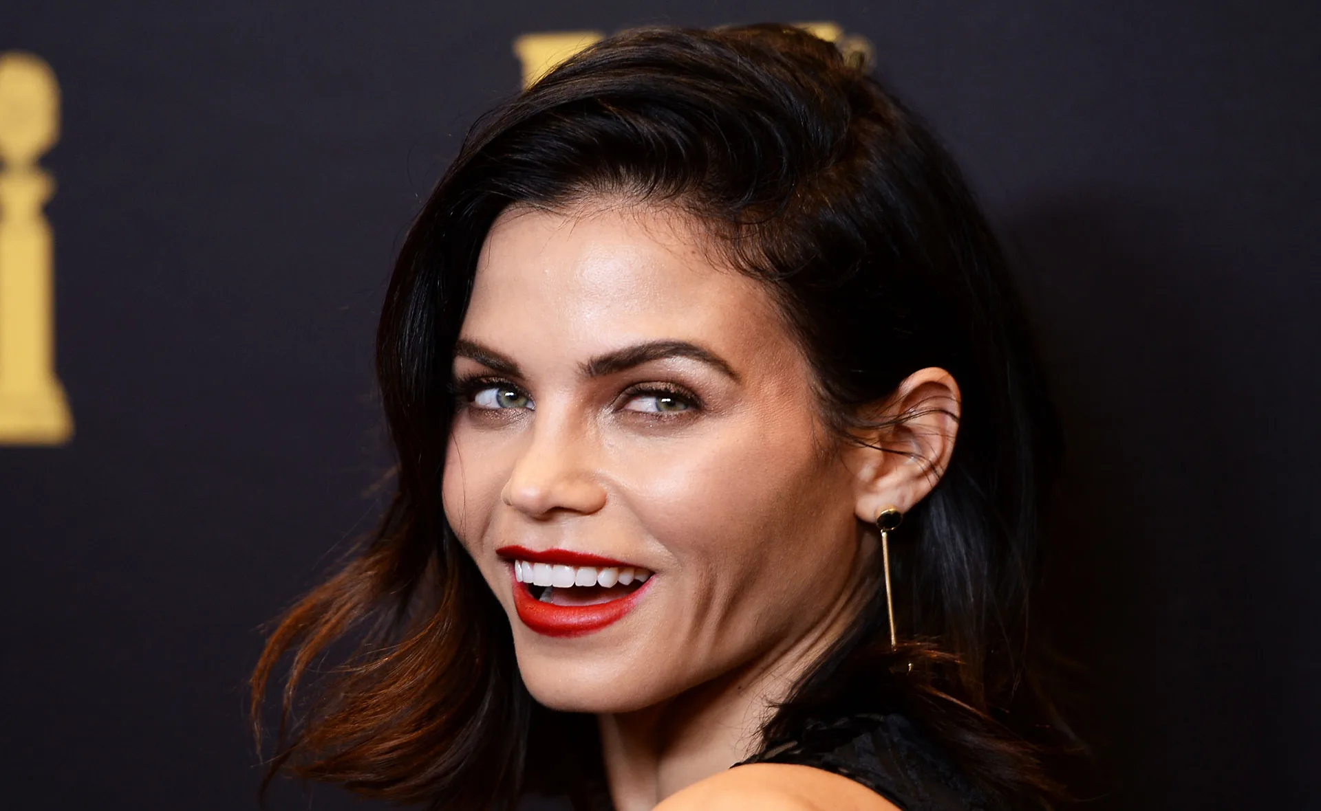 A woman with dark hair and red lipstick smiles, wearing a black outfit and long earrings against a dark background.