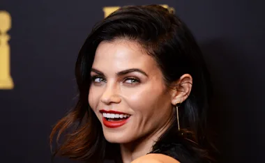 A woman with dark hair and red lipstick smiles, wearing a black outfit and long earrings against a dark background.