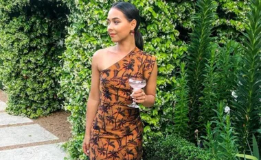 Brooke Blurton in a brown one-shoulder dress with a drink, standing against a lush green background.