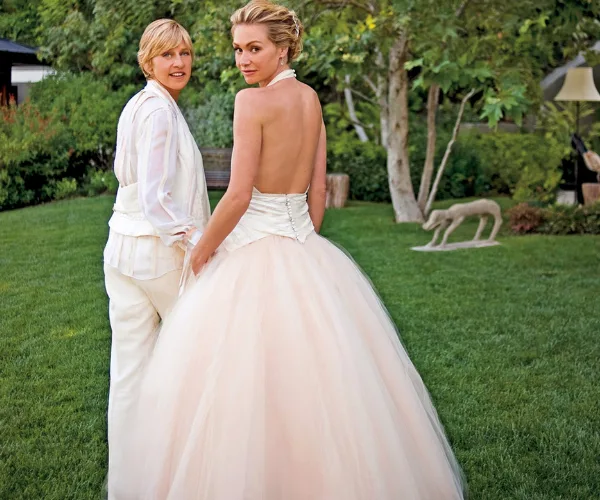 A couple stands in a garden, one in a suit, the other in a strapless wedding gown, both looking back at the camera.