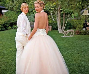 A couple stands in a garden, one in a suit, the other in a strapless wedding gown, both looking back at the camera.