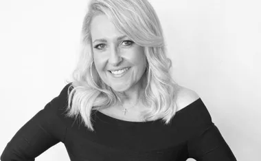 A woman with long blonde hair, smiling, wearing a black off-shoulder top and a cross necklace.