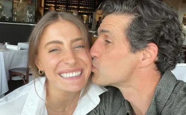 A person smiles broadly while another person kisses their cheek in a cozy restaurant setting.
