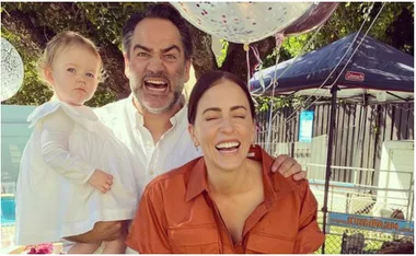 Family smiling with balloons in a backyard, holding a young child in a white dress.