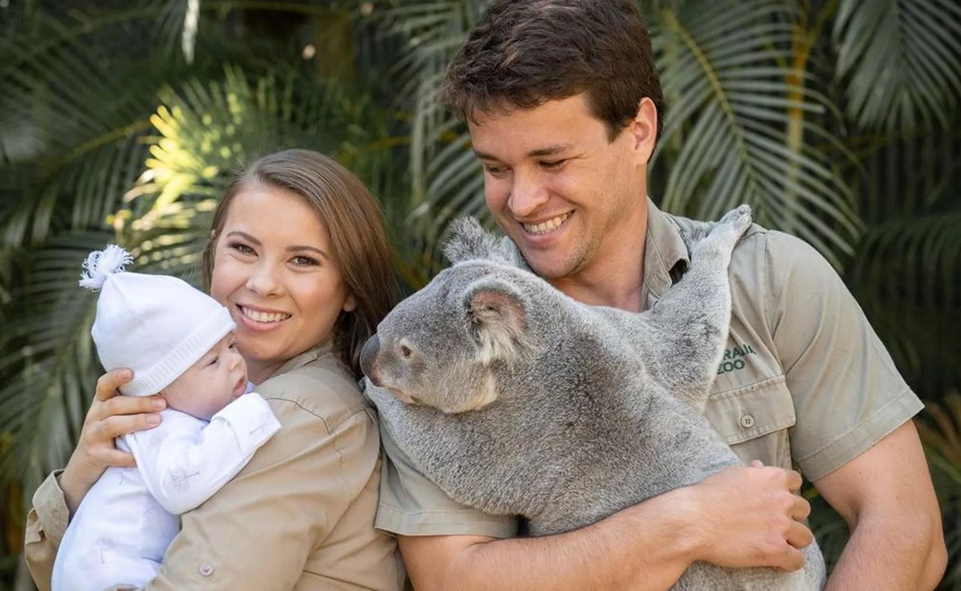 A family holding a baby and a koala, smiling in front of lush green foliage.